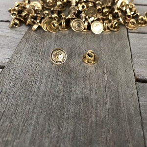 Puede incluir: Un montón de botones de metal de color dorado con un diseño decorativo. Los botones tienen un vástago en la parte posterior para coser.