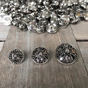 May include: Three silver metal buttons with intricate floral designs. The buttons are arranged in a row from smallest to largest on a wooden surface.