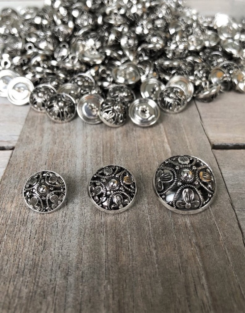 May include: Three silver metal buttons with intricate floral designs. The buttons are arranged in a row from smallest to largest on a wooden surface.