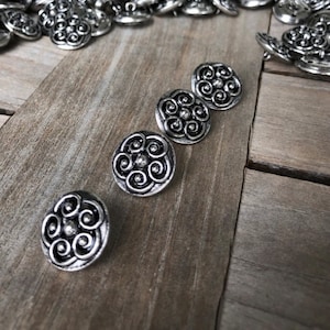 Può includere: Cinque bottoni in metallo argentato con un design a spirale decorativo. I bottoni sono disposti in fila su una superficie di legno.