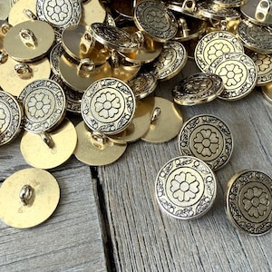 以下が含まれることがあります： 丸い金色のボタンのコレクションで、花の模様が施されています。 ボタンの中央には浮き彫りの花の模様があり、装飾的な縁で囲まれています。 ボタンは木製の表面に散らばっています。