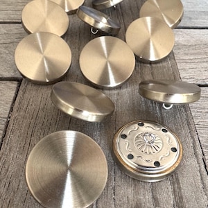 Könnte beinhalten: Eine Sammlung von 11 runden, goldfarbenen Metallknöpfen mit glänzender Oberfläche. Ein Knopf hat ein dekoratives Design.