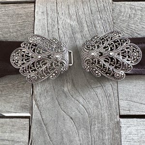 May include: Two silver filigree belt buckles with a decorative floral design. The buckles are on a brown leather belt.