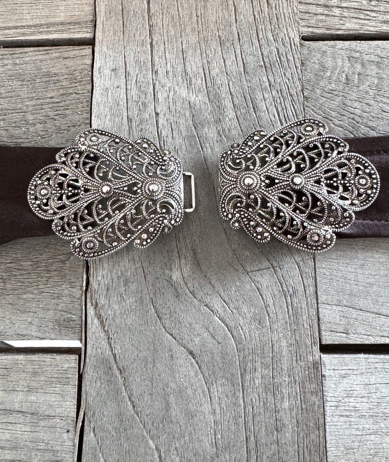 May include: Two silver filigree belt buckles with a decorative floral design. The buckles are on a brown leather belt.