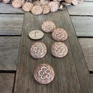 Op de afbeelding: Een verzameling ronde, koperkleurige knopen met een wit bloemenmotief. De knopen hebben een steelrug.