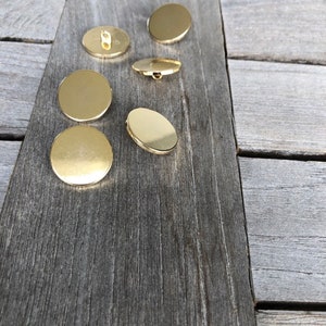 Op de afbeelding: Zes ronde gouden knopen met een glad oppervlak. De knopen zijn in een halve cirkel op een houten oppervlak gerangschikt.