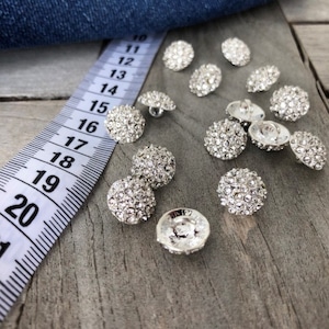 Op de afbeelding: Een close-up van een groep zilverkleurige knopen met heldere strass steentjes. De knopen liggen verspreid op een houten oppervlak naast een witte meetlint.