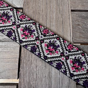 Könnte beinhalten: Ein handgehäkelter Riemen mit einem geometrischen Muster aus schwarzen, weißen, lila und rosa Diamanten. Der Riemen ist diagonal auf einer Holzoberfläche platziert und zeigt die komplizierte Sticharbeit und Farbpalette.