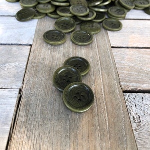 May include: A pile of dark green plastic buttons with four holes each. The buttons are arranged in a pile on a wooden surface.