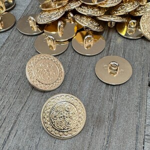 Peut inclure: Un tas de boutons en métal couleur or avec un motif décoratif. Les boutons ont une tige au dos et un motif en relief sur le devant.