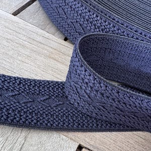 Pode incluir: Close-up de um rolo de acabamento el&aacute;stico tecido azul marinho. O acabamento tem uma superf&iacute;cie texturizada com um padr&atilde;o de diamante repetido. O el&aacute;stico est&aacute; sobre uma superf&iacute;cie de madeira.