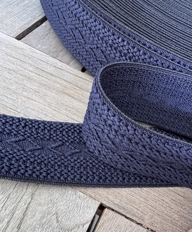 Pode incluir: Close-up de um rolo de acabamento el&aacute;stico tecido azul marinho. O acabamento tem uma superf&iacute;cie texturizada com um padr&atilde;o de diamante repetido. O el&aacute;stico est&aacute; sobre uma superf&iacute;cie de madeira.