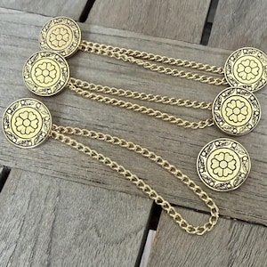 Puede incluir: Cubiertas de botones de metal dorado con un diseño floral, conectadas por delicadas cadenas. Los botones son redondos con un patrón floral detallado en el centro. Las cadenas miden unos 7,6 cm de largo.