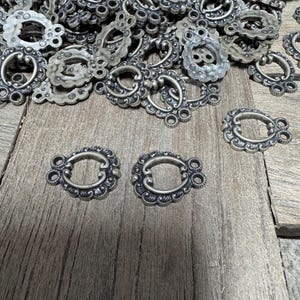 May include: A pile of silver-toned metal decorative clasps with ornate designs. The clasps have a loop on one side and two small rings on the other side.