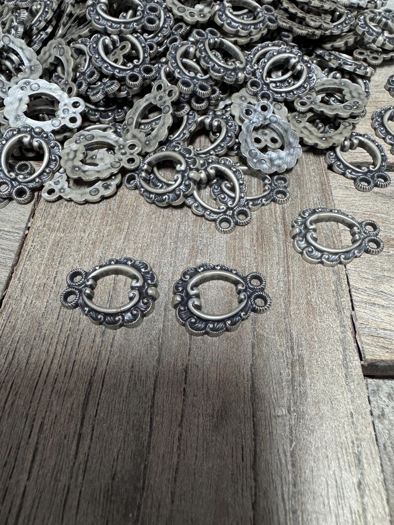 May include: A pile of silver-toned metal decorative clasps with ornate designs. The clasps have a loop on one side and two small rings on the other side.