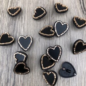 May include: A collection of black heart-shaped buttons with gold-colored edges. The buttons have a textured, slightly raised design around the edges.