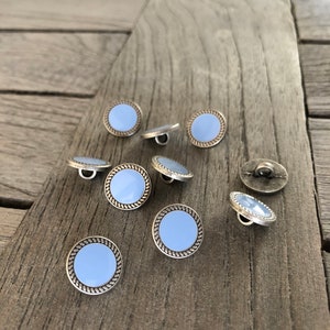 Puede incluir: Ocho botones redondos de color azul claro con bordes de metal plateado y un diseño de cuerda. Los botones tienen dos agujeros cada uno.