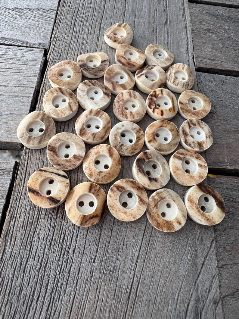 May include: A collection of round buttons with two holes each. The buttons have a light tan base color with brown and cream-colored marbling. The buttons are arranged on a weathered wooden surface.
