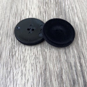 May include: Two black buttons, one with a smooth surface and the other with a textured surface and multiple holes.
