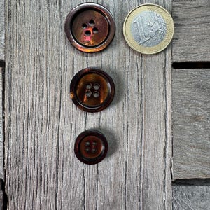 May include: Three brown buttons with a pearlescent sheen, each with a different size and number of holes. A one Euro coin is next to the largest button. The buttons are on a weathered, gray wooden surface.