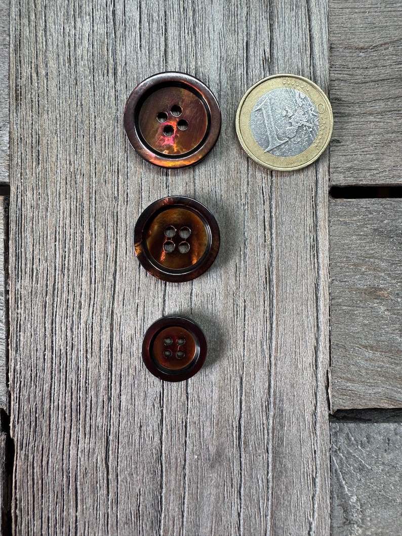 May include: Three brown buttons with a pearlescent sheen, each with a different size and number of holes. A one Euro coin is next to the largest button. The buttons are on a weathered, gray wooden surface.