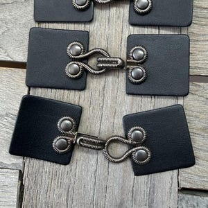 Puede incluir: Tres juegos de cierres de palanca de cuero negro con herrajes metálicos plateados. Cada cierre presenta una base cuadrada de cuero con detalles metálicos decorativos y un cierre de bucle. Los cierres están dispuestos sobre una superficie de madera envejecida.