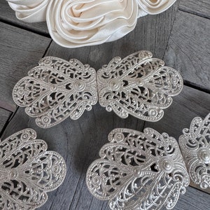 K&ouml;nnte beinhalten: Vier silberne filigranierte G&uuml;rtelschnallen aus Metall mit einem dekorativen Blumenmuster. Die Schnallen sind in einer Reihe auf einer Holzoberfl&auml;che angeordnet.