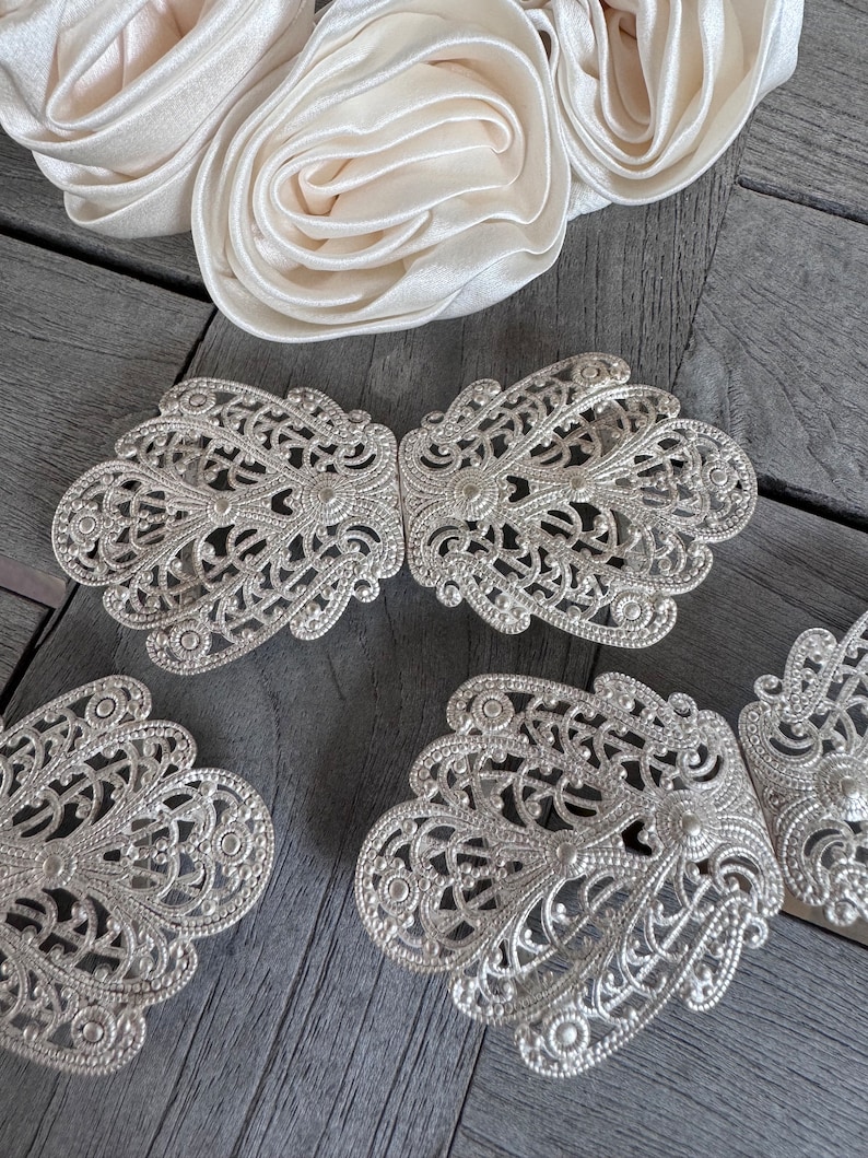 K&ouml;nnte beinhalten: Vier silberne filigranierte G&uuml;rtelschnallen aus Metall mit einem dekorativen Blumenmuster. Die Schnallen sind in einer Reihe auf einer Holzoberfl&auml;che angeordnet.