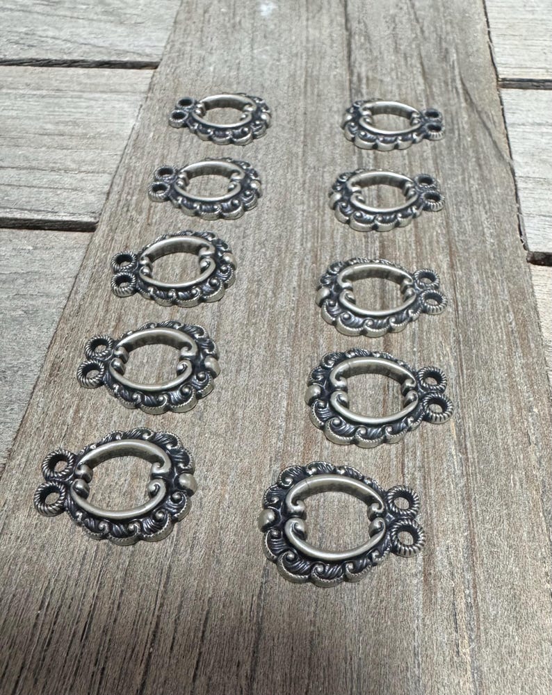 May include: Ten silver-toned metal decorative embellishments with a scalloped oval design. Each embellishment has two loops on the top.