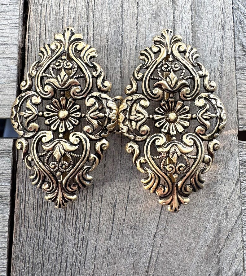 May include: Ornate gold-colored metal clasp with an intricate floral and scroll design. The clasp is composed of two symmetrical halves connected by a hinge. The design includes stylized flowers and swirling patterns, creating a vintage aesthetic.
