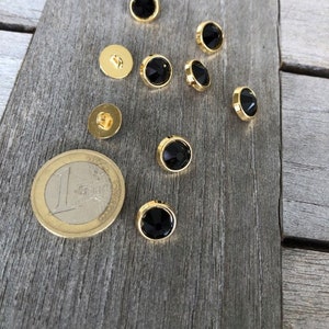 May include: Eight gold-toned buttons with black glass centres. The buttons are arranged in a semi-circle on a wooden surface.