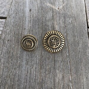 May include: Two gold-toned metal buttons with intricate designs. The larger button has a circular design with a floral pattern in the center. The smaller button has a similar design but is smaller in size.