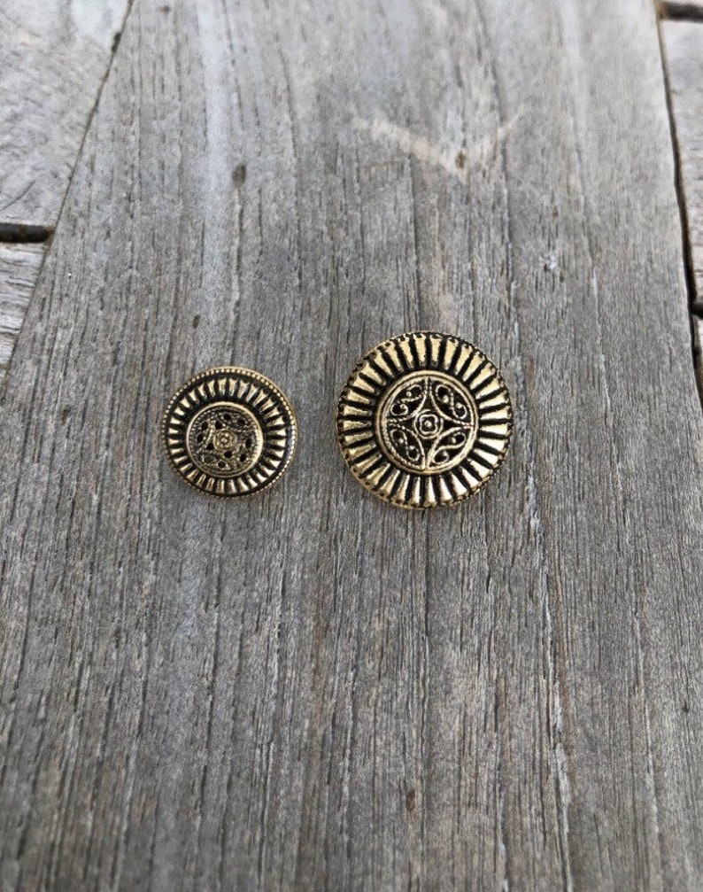 May include: Two gold-toned metal buttons with intricate designs. The larger button has a circular design with a floral pattern in the center. The smaller button has a similar design but is smaller in size.