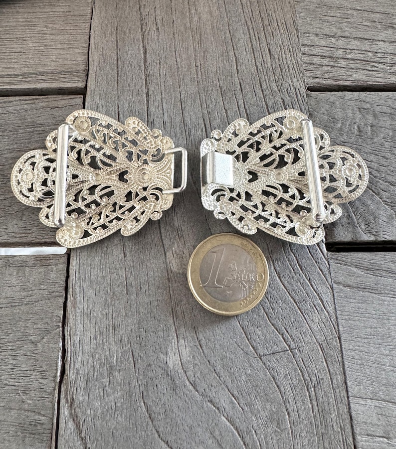 May include: Two silver filigree belt buckles with a decorative floral design. The buckles are about the size of a Euro coin.