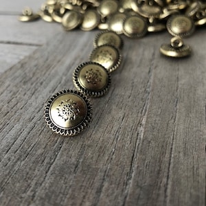 May include: A pile of gold-toned metal buttons with a decorative design. The buttons have a raised edge and a floral pattern in the center.
