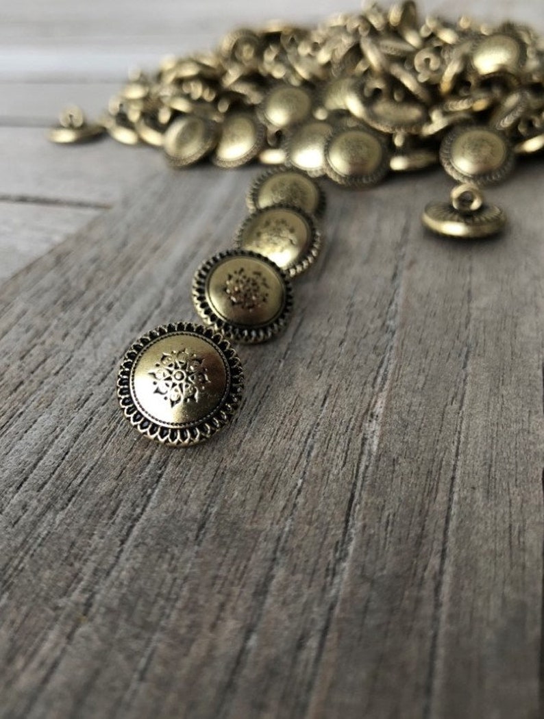 May include: A pile of gold-toned metal buttons with a decorative design. The buttons have a raised edge and a floral pattern in the center.