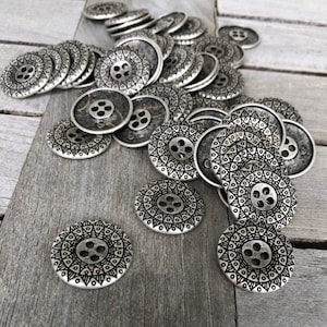 Può includere: Un mucchio di bottoni in metallo argentato con un design decorativo a mandala. I bottoni hanno quattro fori e sono disposti in modo casuale.