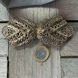 May include: Ornate antique gold-tone metal clasp with intricate filigree detailing. The clasp is in two symmetrical halves, designed to connect. A Euro coin is next to the clasp for size reference. The clasp is on a weathered wooden surface.