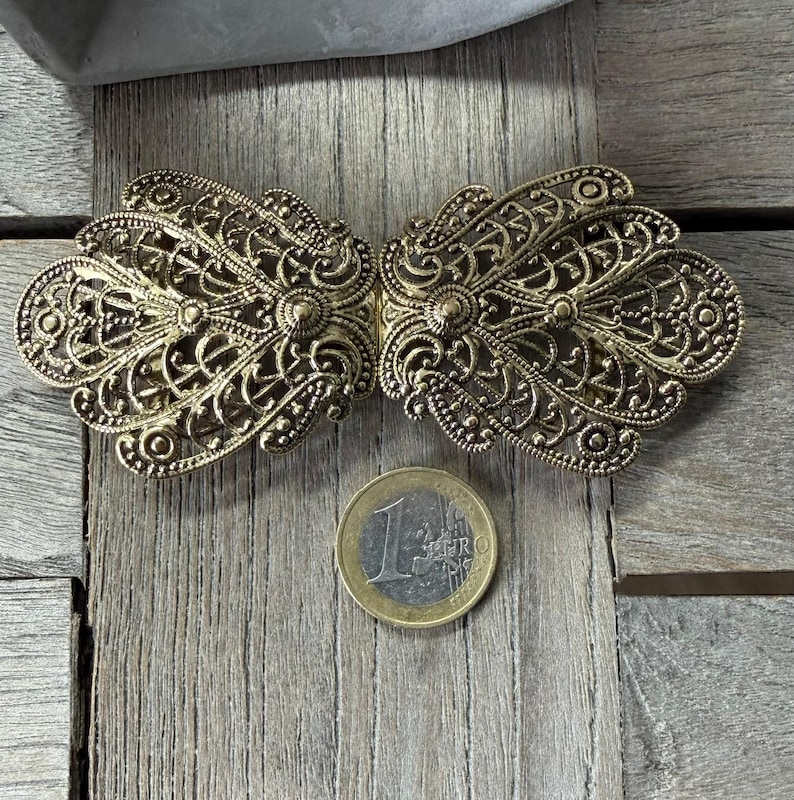 May include: Ornate antique gold-tone metal clasp with intricate filigree detailing. The clasp is in two symmetrical halves, designed to connect. A Euro coin is next to the clasp for size reference. The clasp is on a weathered wooden surface.