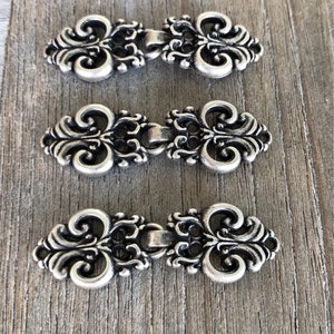 May include: Three silver-toned metal clasps with ornate, scroll-like designs. The clasps are arranged in a row, with the top clasp slightly higher than the others.
