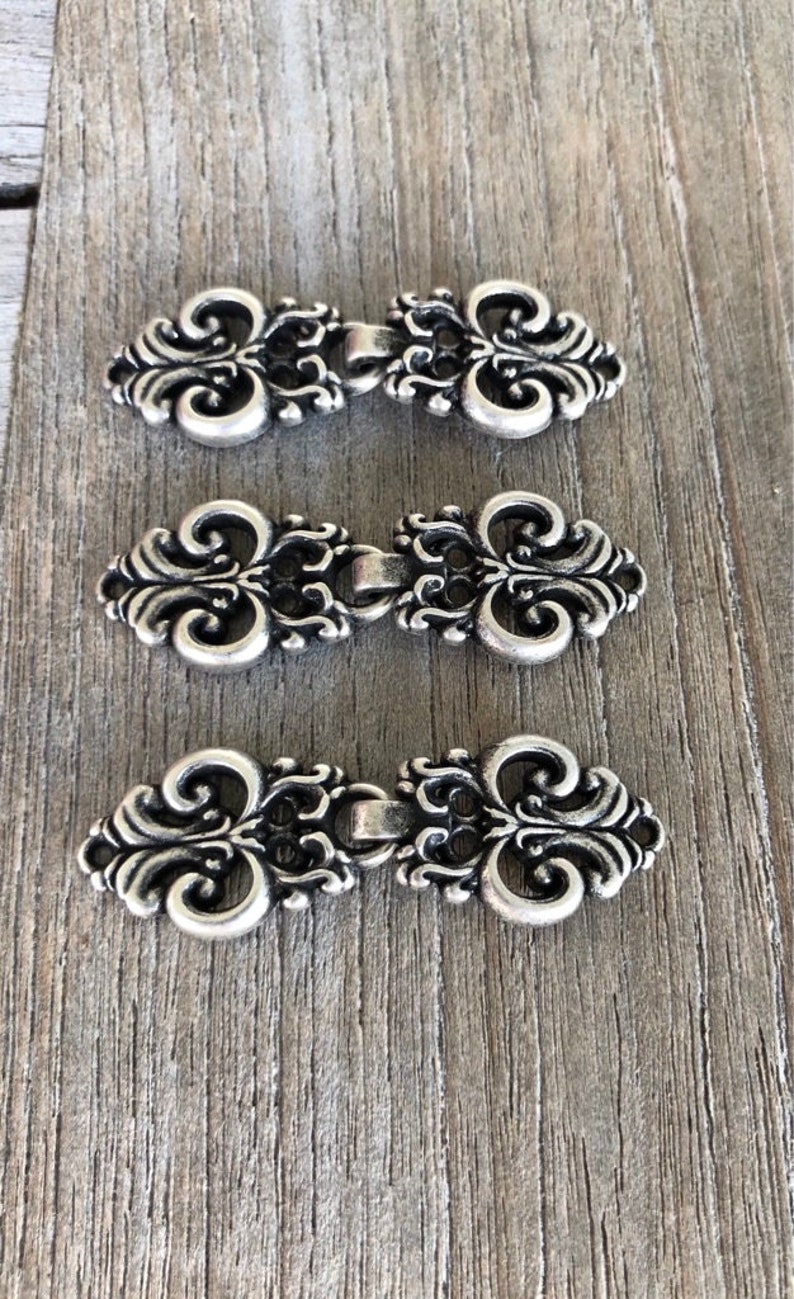 May include: Three silver-toned metal clasps with ornate, scroll-like designs. The clasps are arranged in a row, with the top clasp slightly higher than the others.