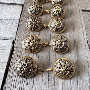 May include: Several antique gold-coloured buttons with detailed circular patterns. Each button is linked to another by a small loop and bar. The buttons are arranged in rows on a weathered wooden surface.
