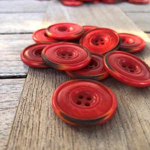 Peut inclure: Un tas de boutons en plastique rouge avec quatre trous chacun. Les boutons ont un bord noir et un effet marbré.