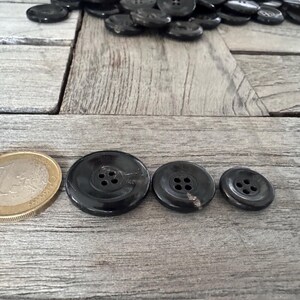 May include: Three black buttons with four holes each. The buttons are arranged in a row from largest to smallest. The largest button is about the size of a UK 20 pence coin.