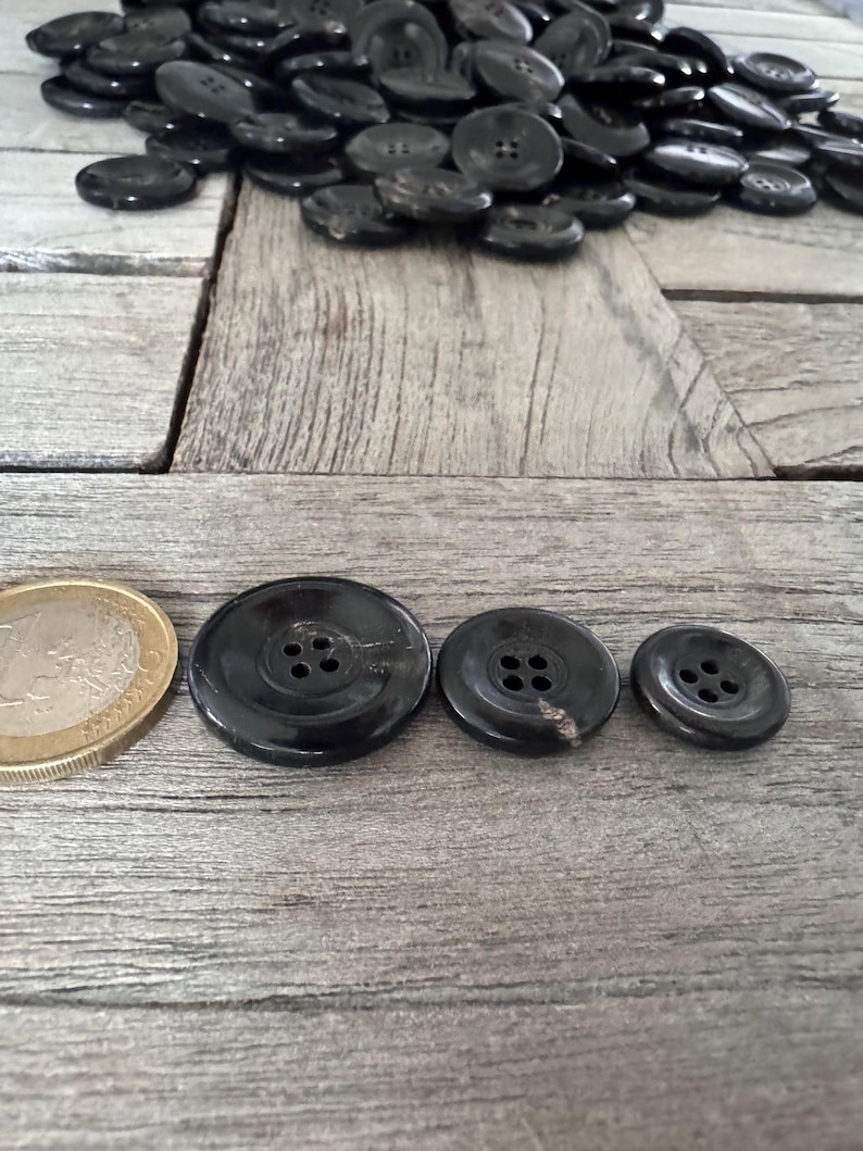 May include: Three black buttons with four holes each. The buttons are arranged in a row from largest to smallest. The largest button is about the size of a UK 20 pence coin.