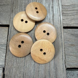 Op de afbeelding: Vijf ronde, lichtbruine houten knopen met elk twee gaten. Ze liggen op een verweerd, grijs houten oppervlak. De knopen hebben een glad, gepolijst uiterlijk en variëren enigszins in kleur.