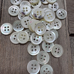 Puede incluir: Un conjunto de botones redondos de color blanco roto con cuatro agujeros, dispersos sobre una superficie de madera. Algunos botones tienen un brillo sutil e iridiscente, mientras que otros presentan un diseño tenue. Los botones varían ligeramente de tamaño.