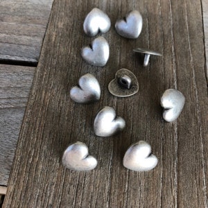 May include: A collection of nine silver heart-shaped buttons. The buttons are arranged on a wooden surface. Some of the buttons have a raised design.