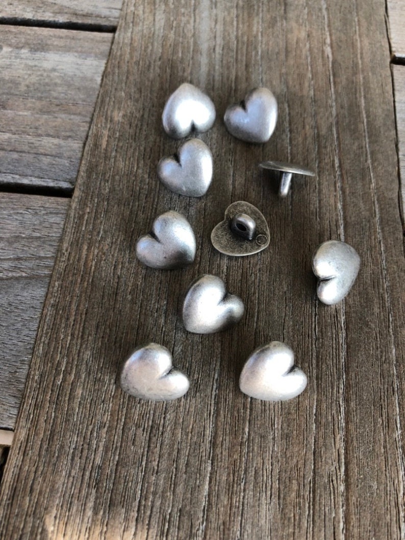 May include: A collection of nine silver heart-shaped buttons. The buttons are arranged on a wooden surface. Some of the buttons have a raised design.
