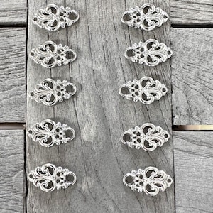 Könnte beinhalten: Acht silberfarbene, dekorative Metallverzierungen mit filigranem Design. Die Verzierungen sind oval und haben jeweils zwei Löcher an den Seiten.